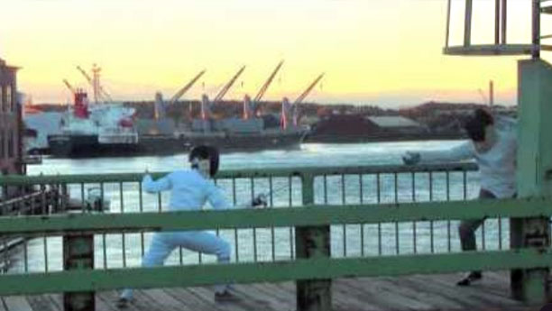 Two fencers duel on a bridge at sunset.